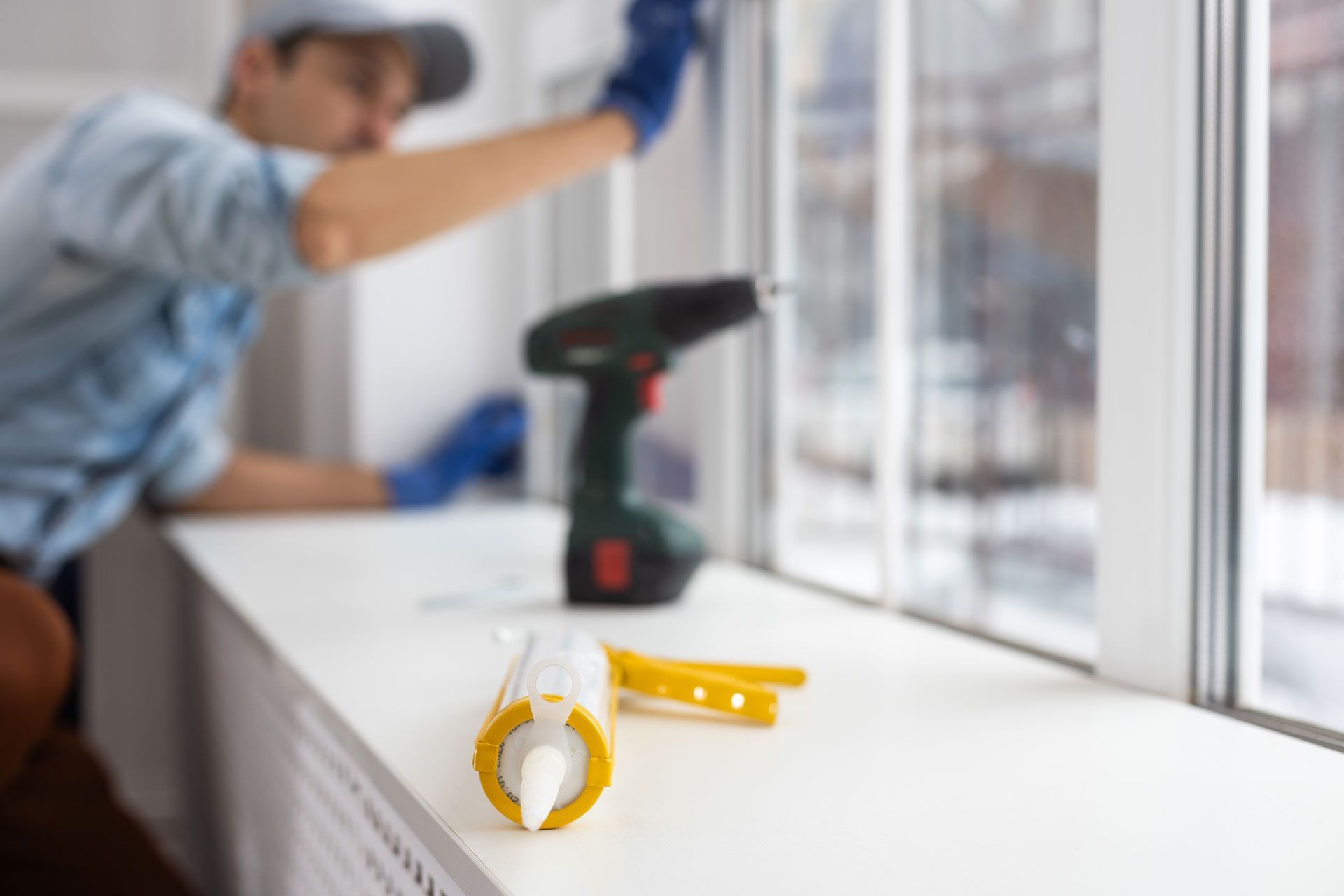 A worker in blue gloves applies sealant to a window frame, with a caulk gun and drill on the sill nearby.
