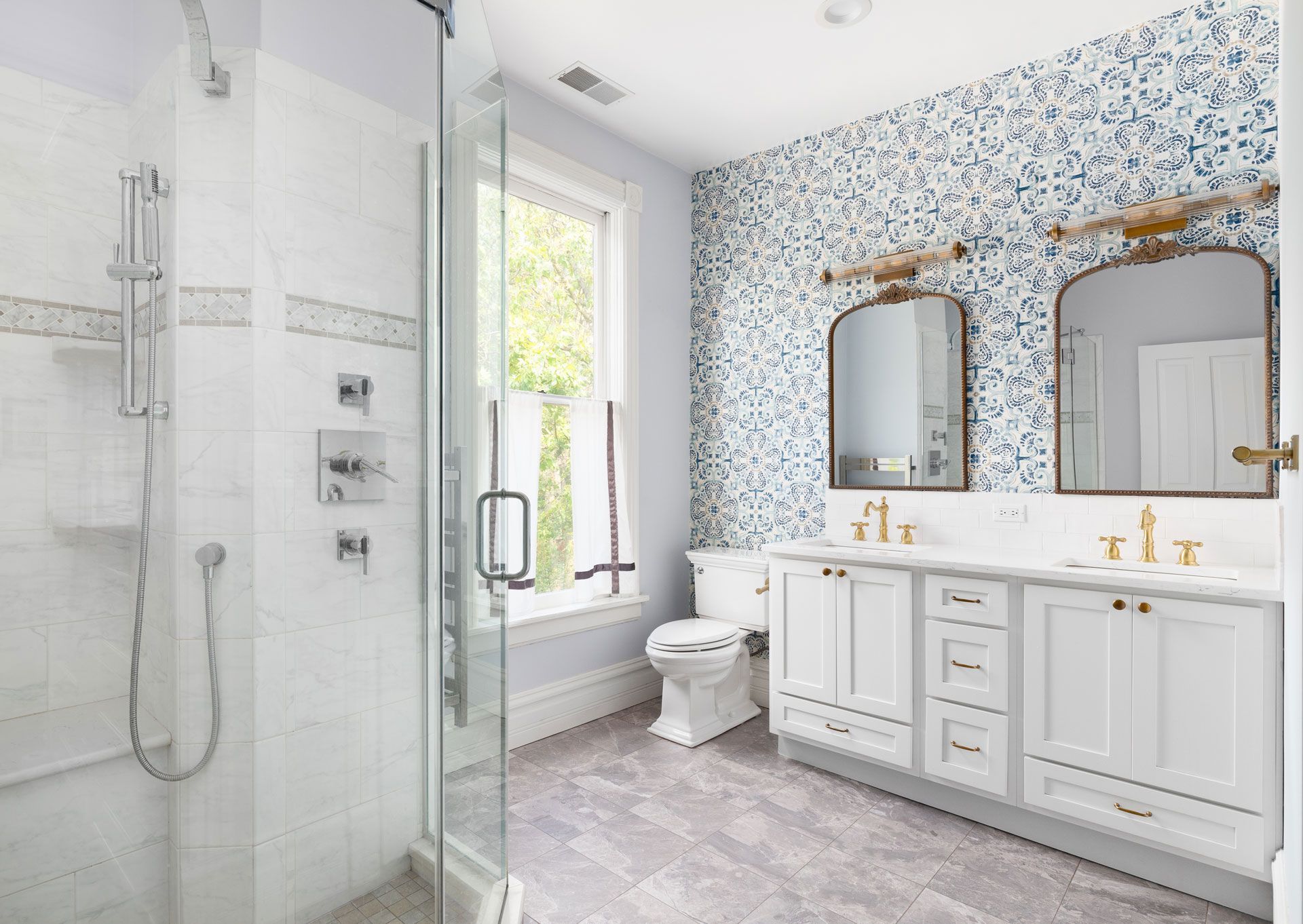 A bright, modern bathroom featuring a white vanity with double sinks, patterned blue wallpaper, and a glass shower stall.
