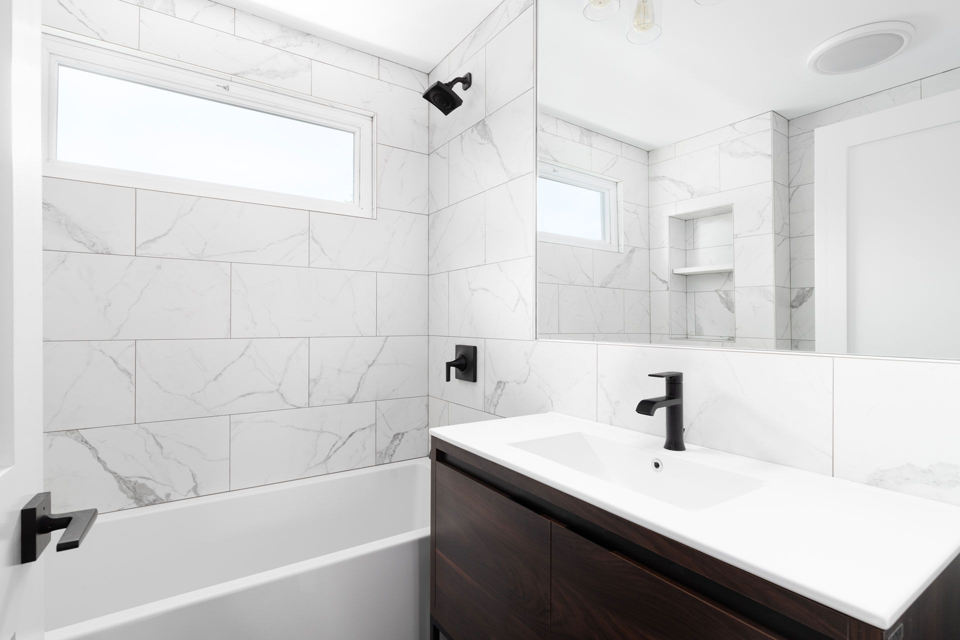 Modern bathroom featuring white marble-look tiled walls, a dark wood vanity, matte black fixtures, and a tub-shower area.