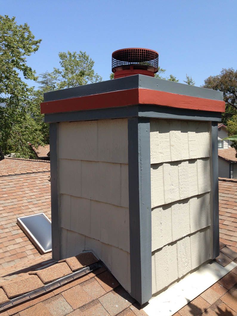 A square, beige-sided chimney with a red-accented cap and dark grey trim, sitting on a brown shingled roof under blue sky.