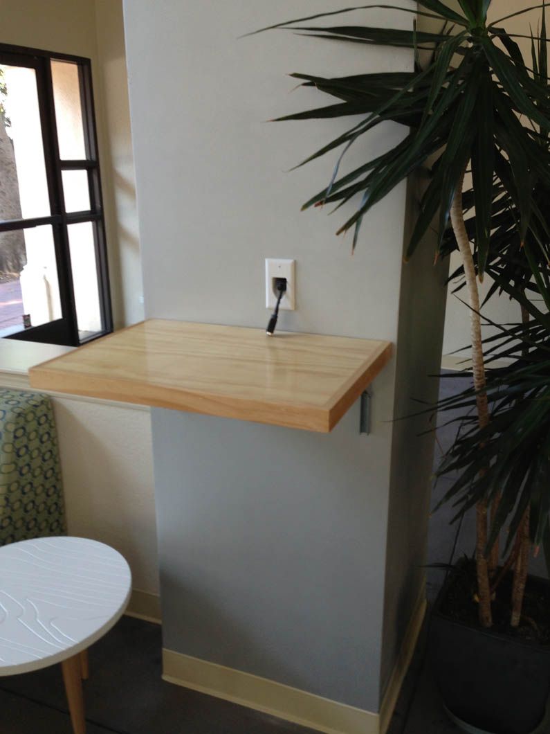 A wooden floating desk mounted on a gray wall with an electrical outlet above it, located next to a potted plant.