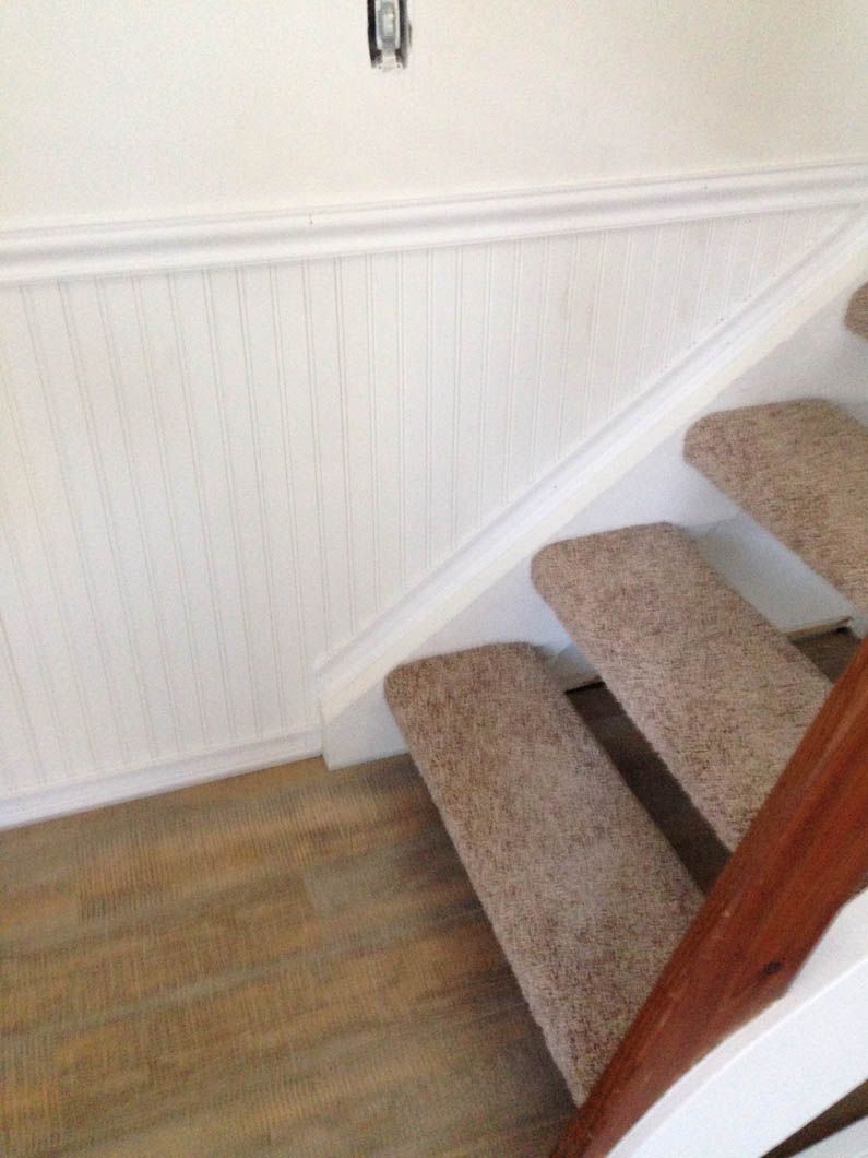 A staircase with tan carpet, a white wainscoted wall, and wood-patterned flooring.