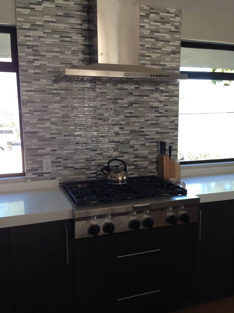 A stainless steel gas stove and range hood set against a grey, horizontal tile backsplash in a modern kitchen.