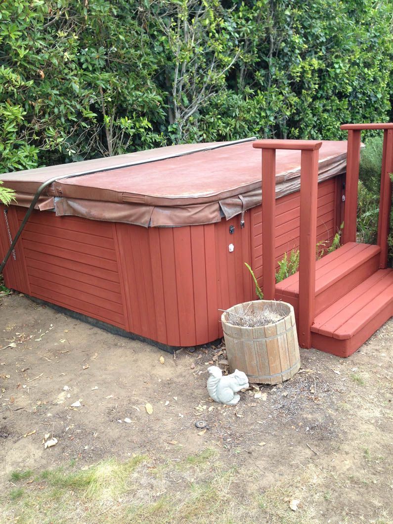 A red hot tub with a brown cover, matching stairs, and a small squirrel statue sits on a dirt patch near green bushes.