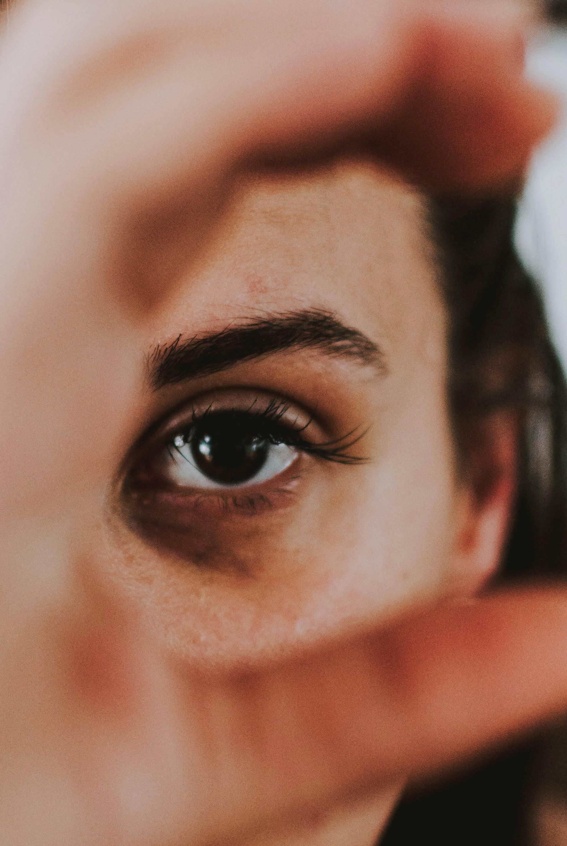 Close-up of a person's eye framed by a hand, brown eye, dark eyebrows and long eyelashes, skin is visible.