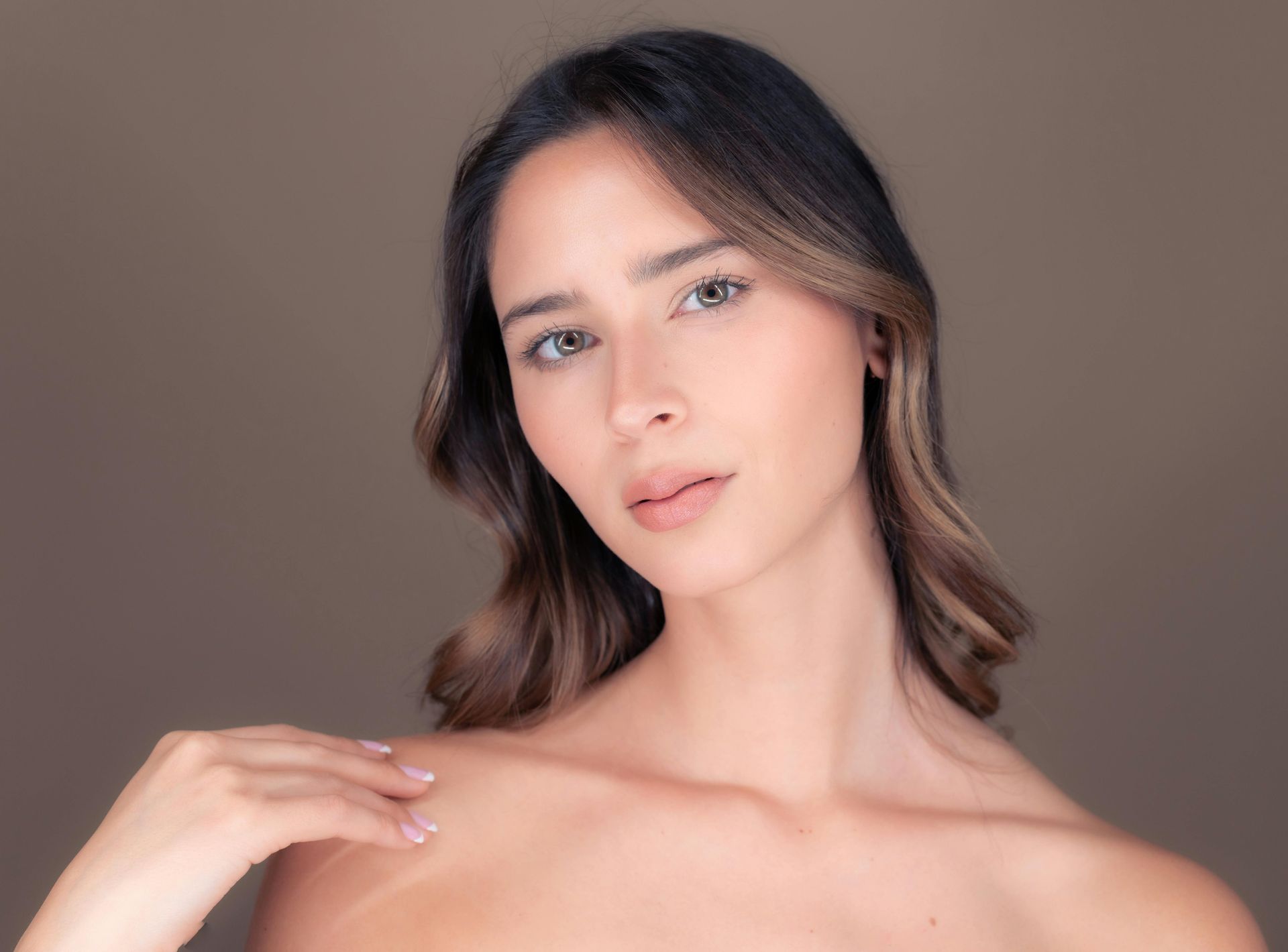 Woman with brown hair and skin, looking at the camera, hand on shoulder, neutral background.