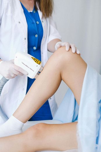 Doctor performing laser hair removal on a patient's leg. Light skin, white coat, blue shirt, gloves.