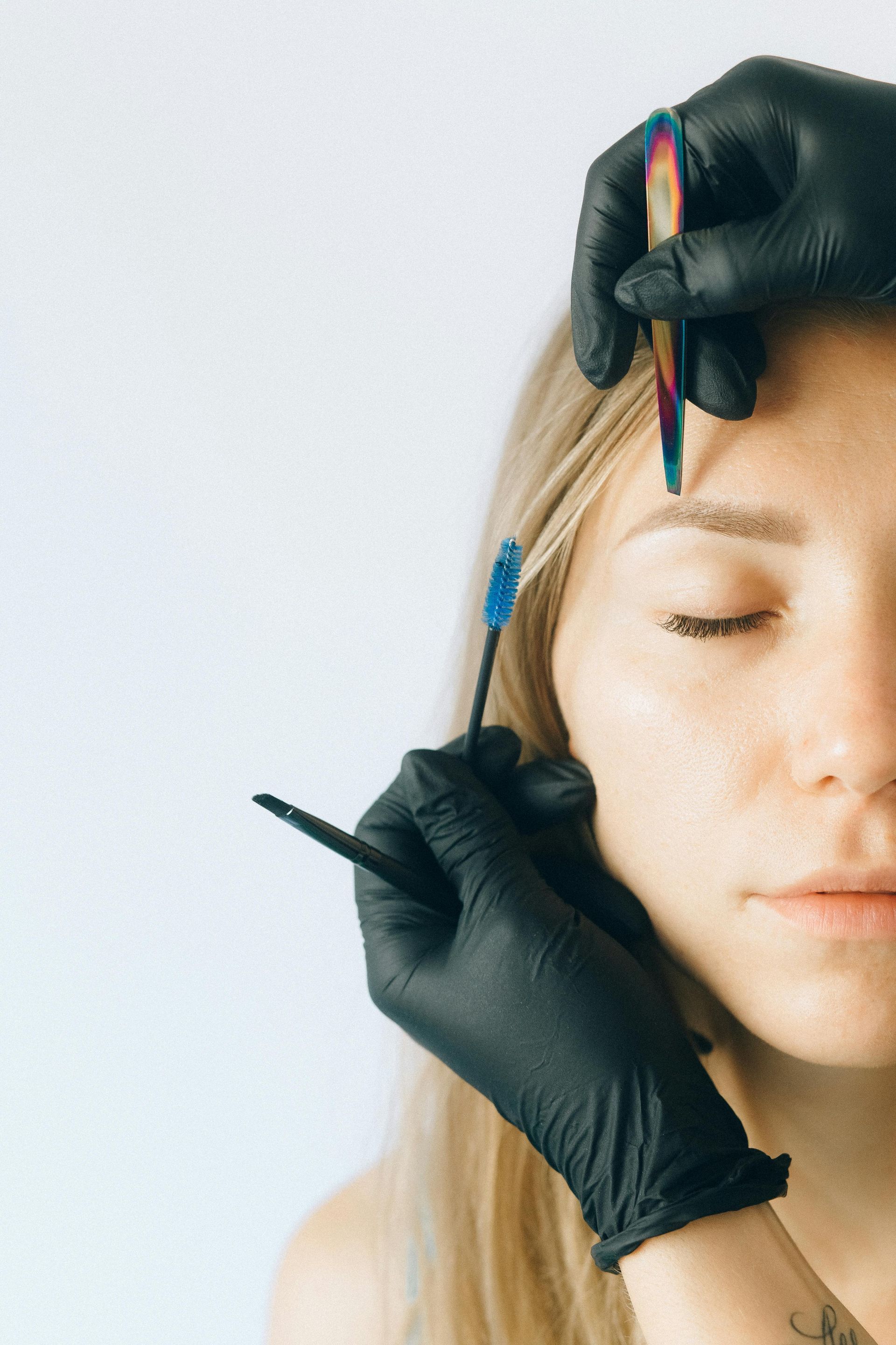 A woman with closed eyes has her eyebrows being groomed by someone wearing black gloves.