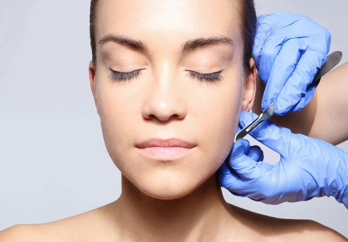 Woman's ear being pierced with surgical tools. Gloved hands, closed eyes, neutral background.