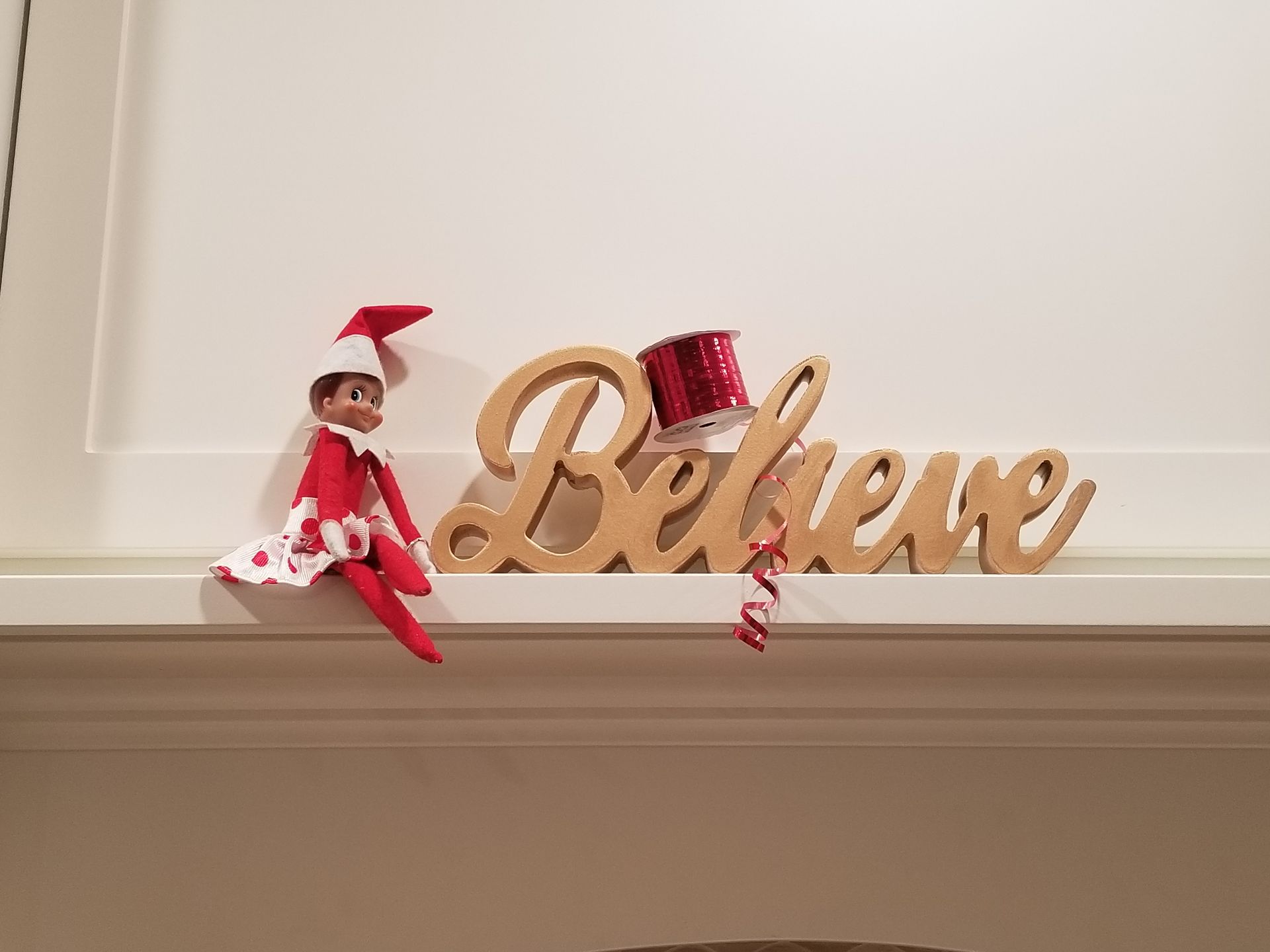 An elf on the shelf is sitting on a shelf next to a wooden sign that says believe.