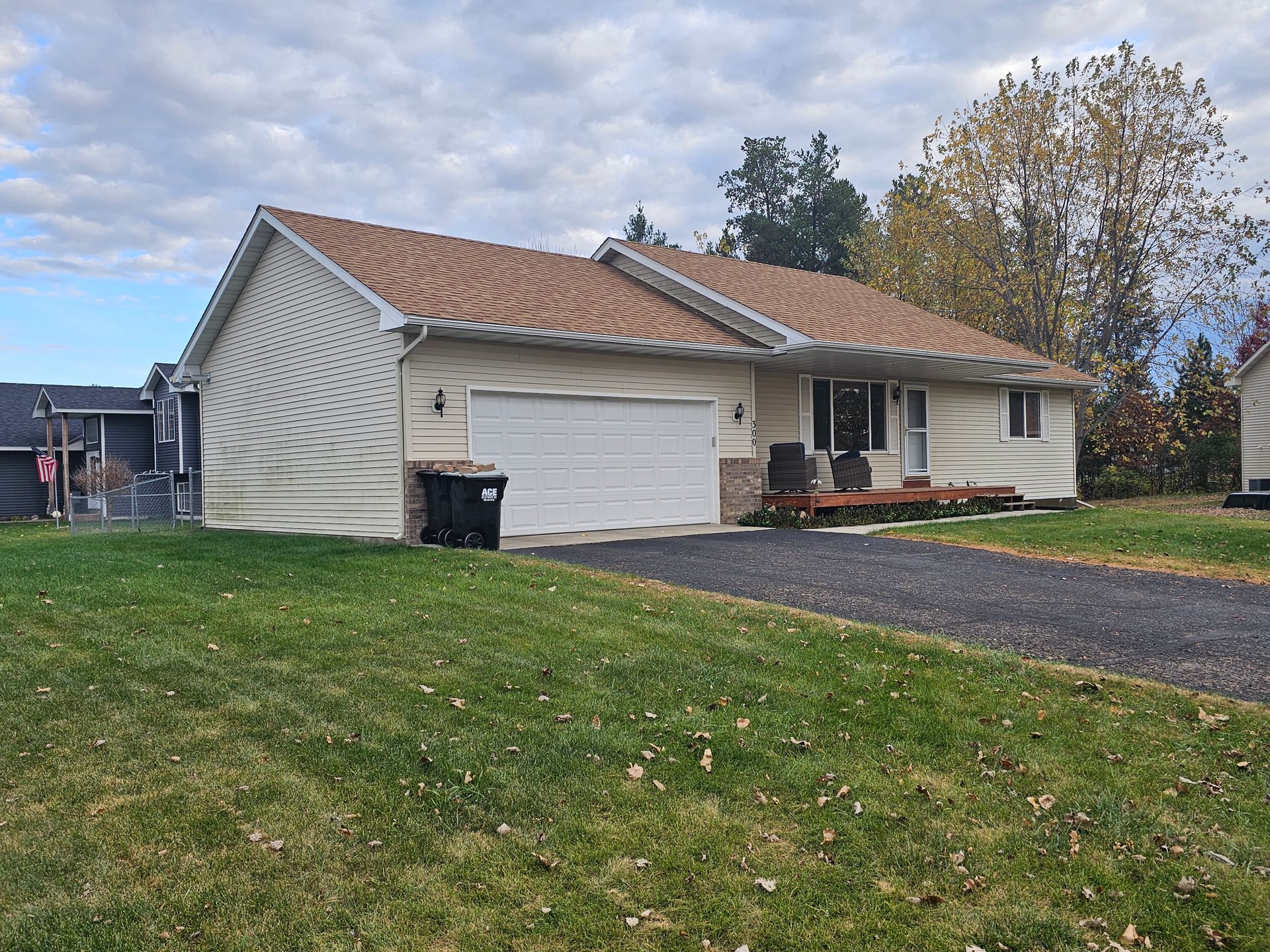 A light-colored, single-story ranch home with a two-car garage, a gravel driveway, and a small front deck on a lawn.
