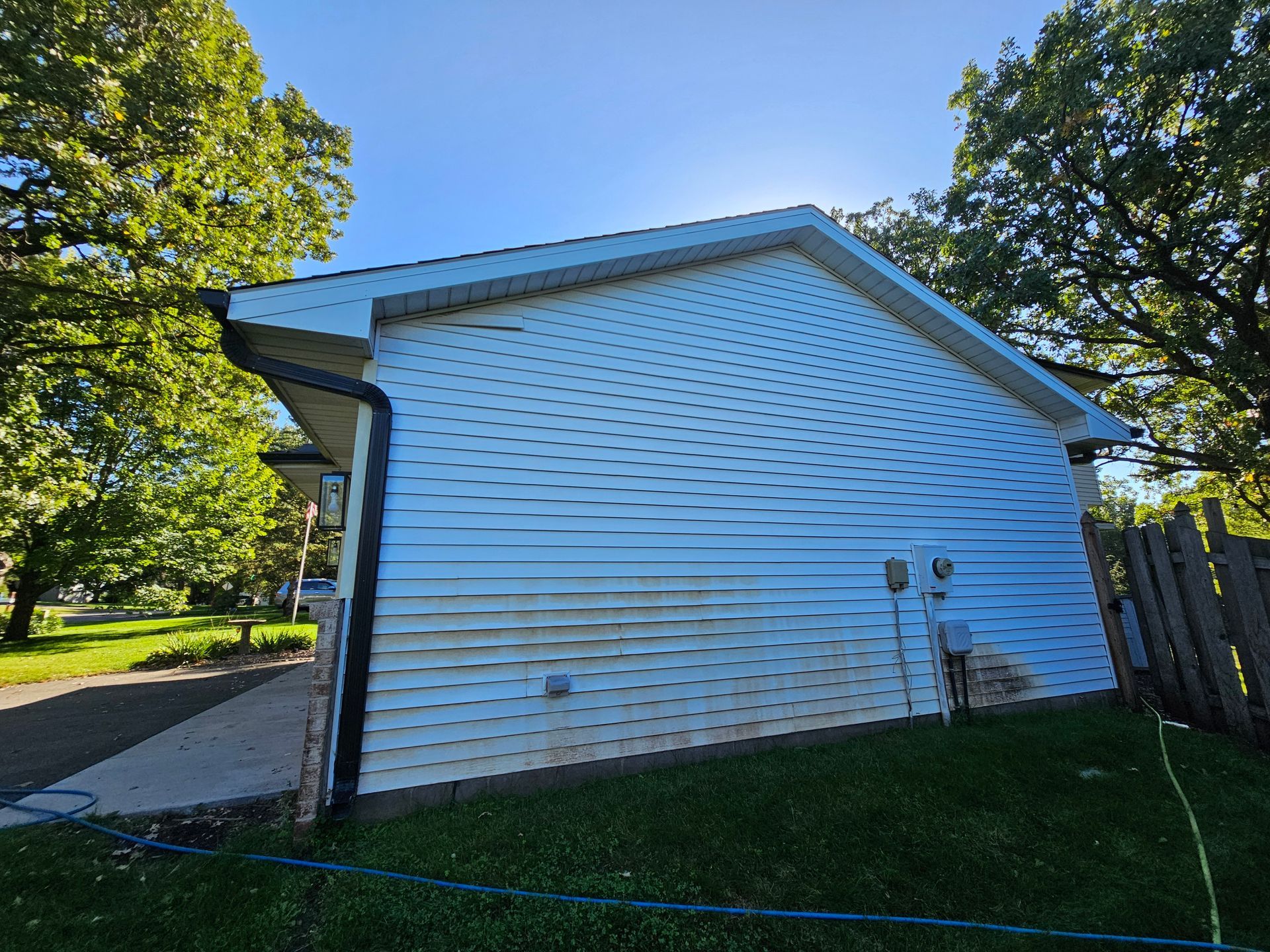Side view of a house with white horizontal siding and a dark roof, surrounded by green trees and a lawn.