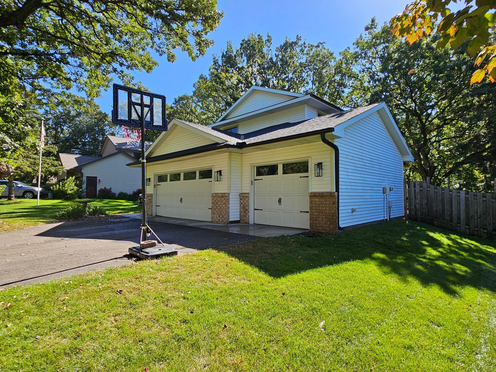 A two-car garage with white siding and stone accents sits on a residential lot with a portable basketball hoop in front.