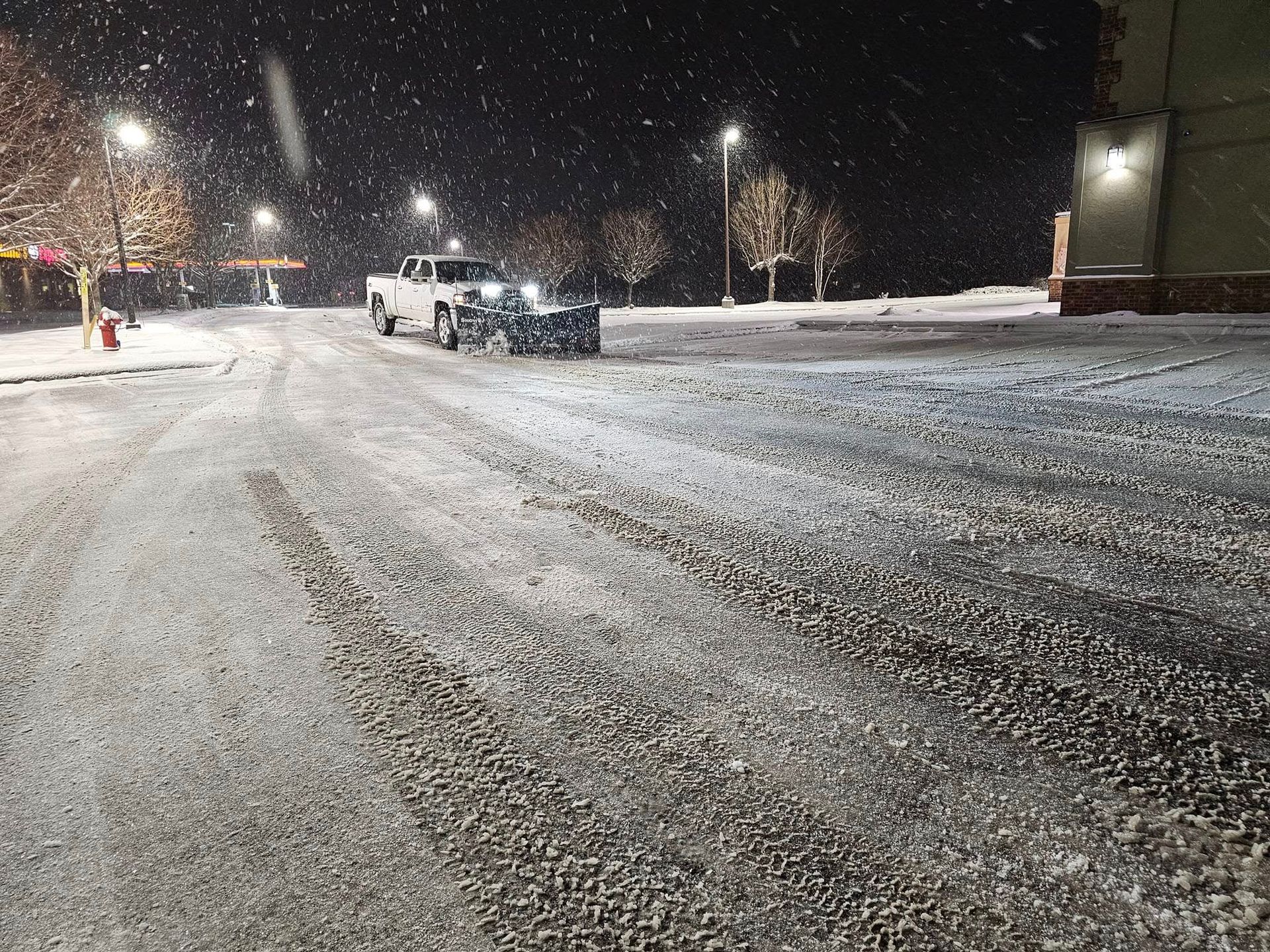 Snow-covered road at night, truck pulling trailer driving. Streetlights illuminate falling snow.