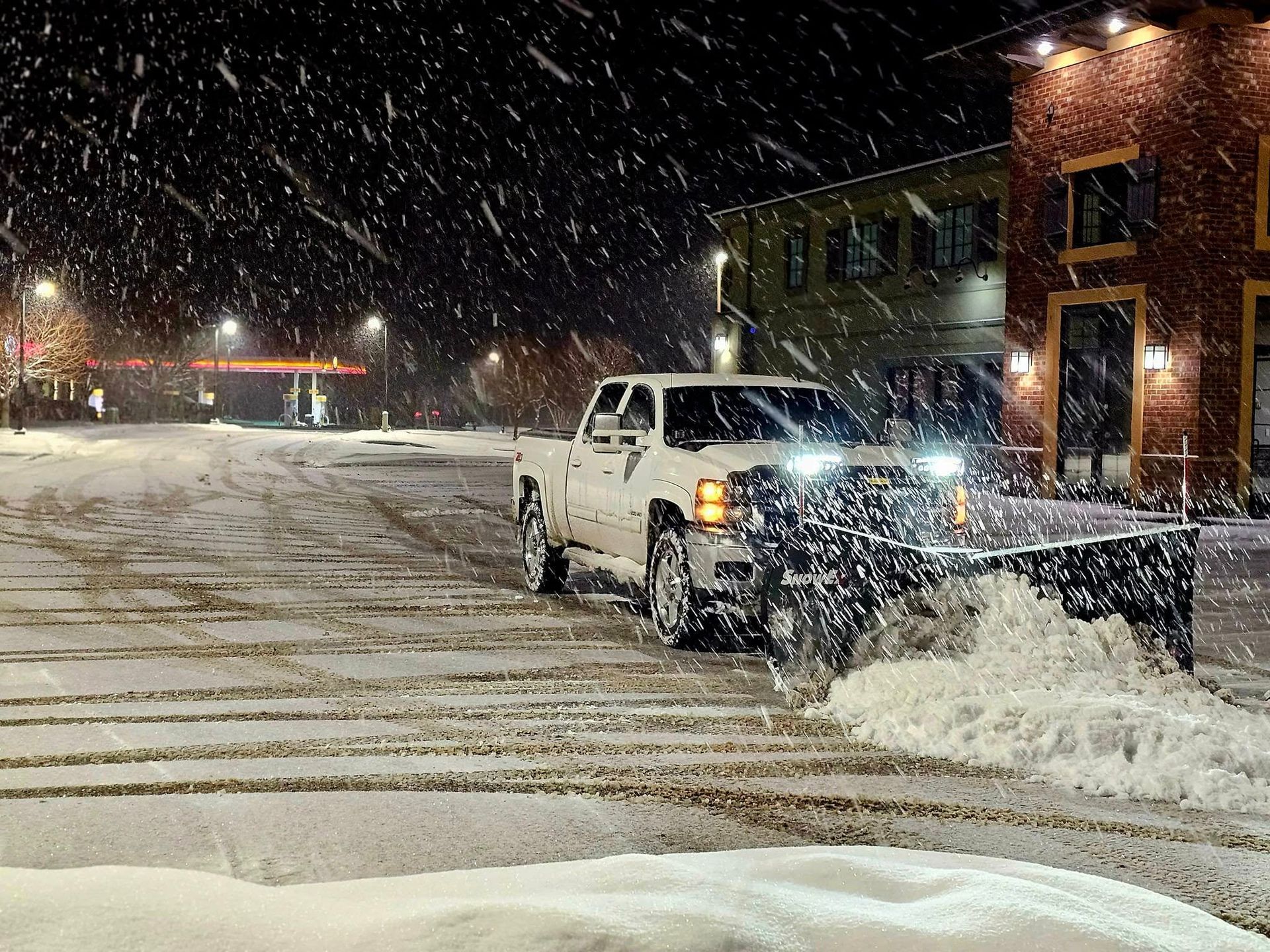 Snowplow clearing snow from a street at night, with buildings and a gas station visible.