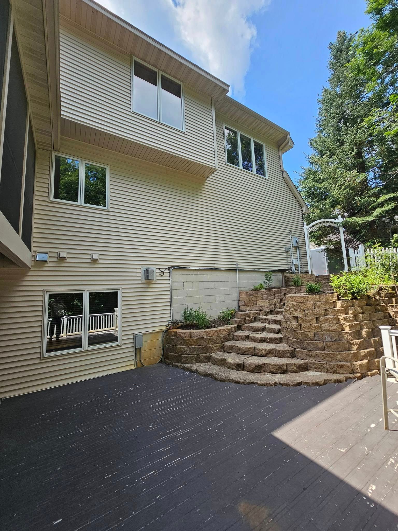 Exterior of a two-story house with a wooden deck, stone steps, and surrounding trees under a blue sky.