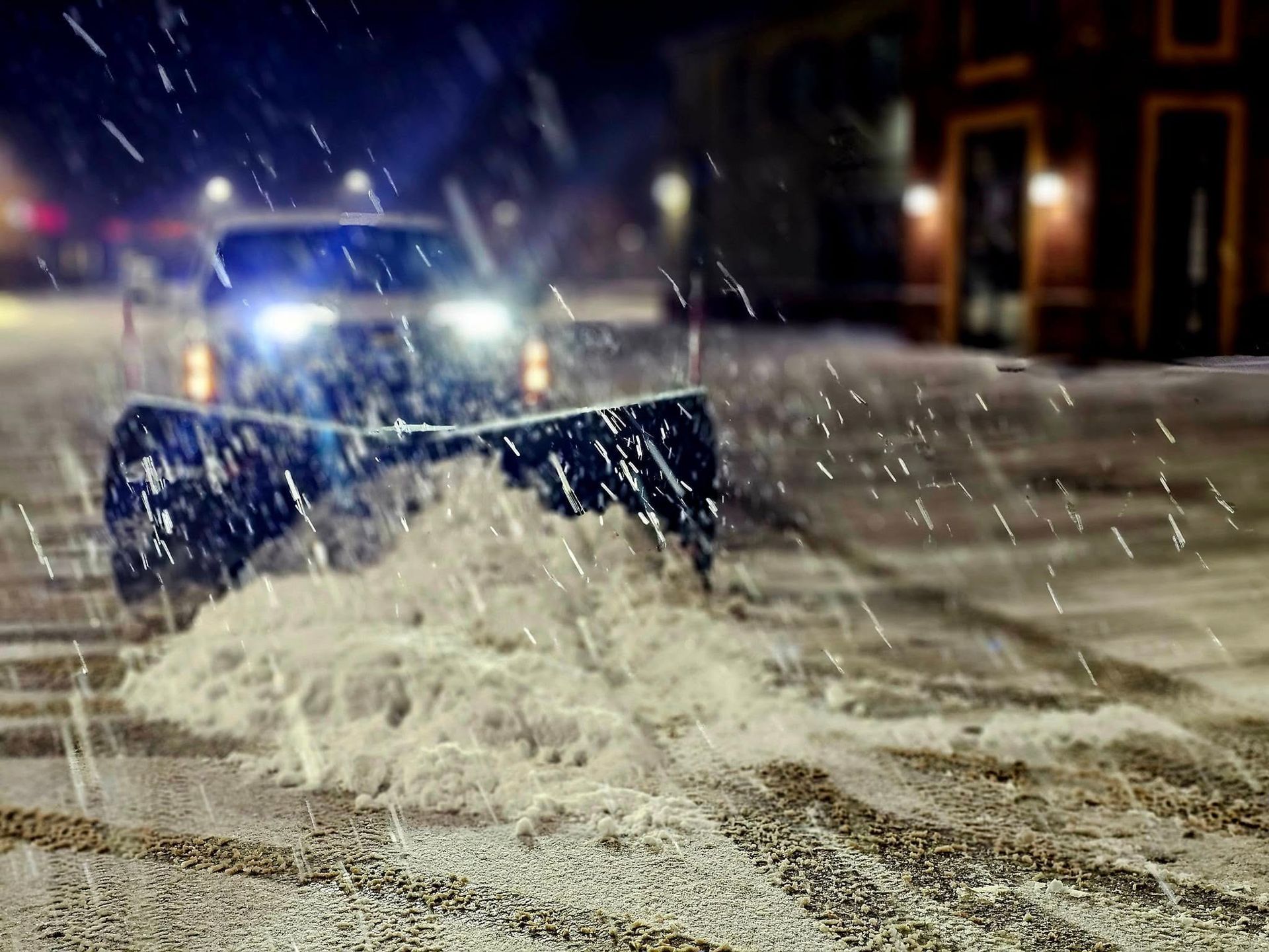 Snowplow clearing snow from a street at night; headlights visible, snow falling.