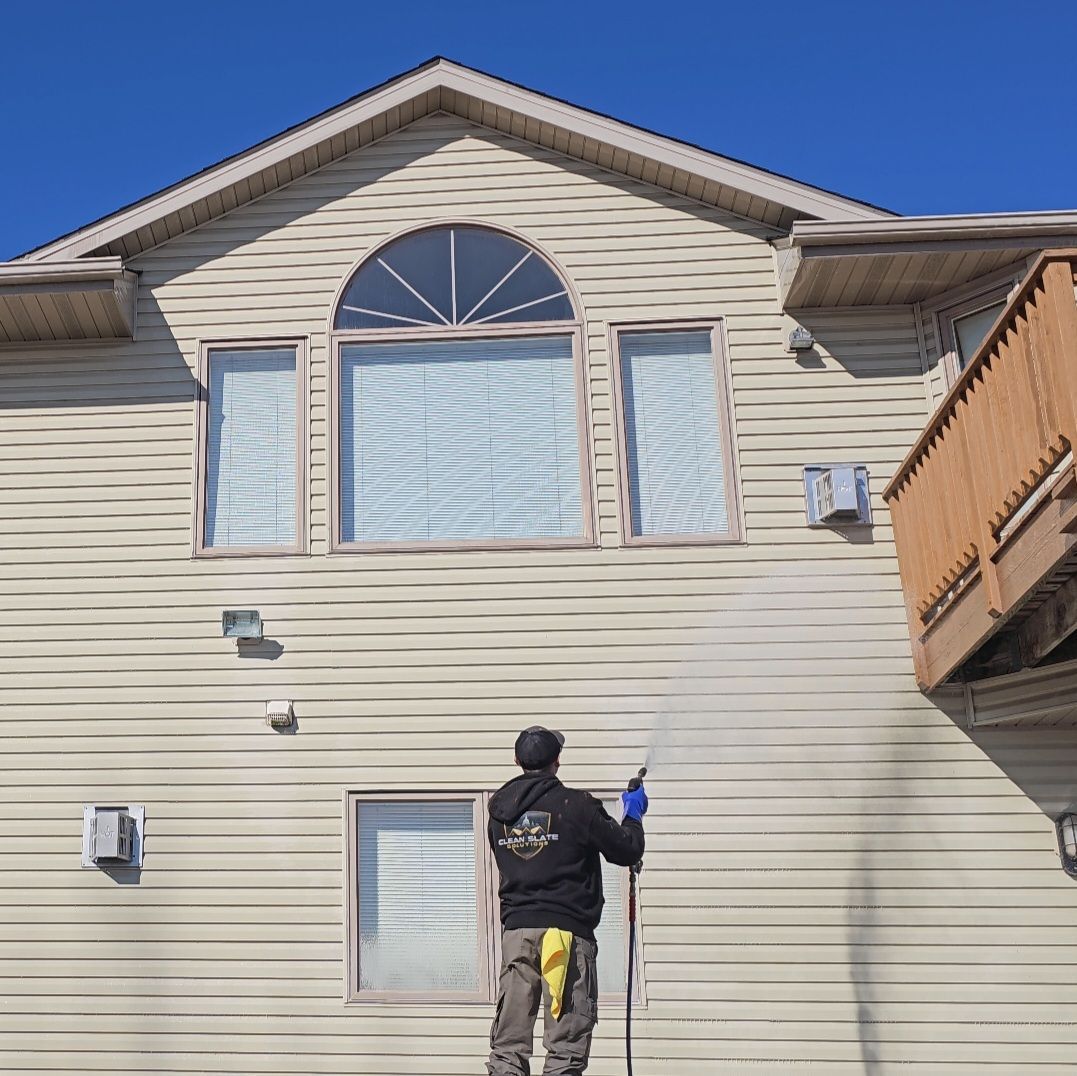 A person in a black jacket power washes the light-colored vinyl siding of a two-story residential home.