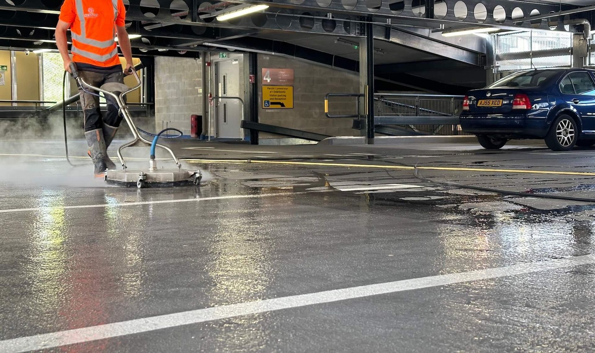Person in orange vest using a floor cleaning machine on a wet paved area near a parked car.