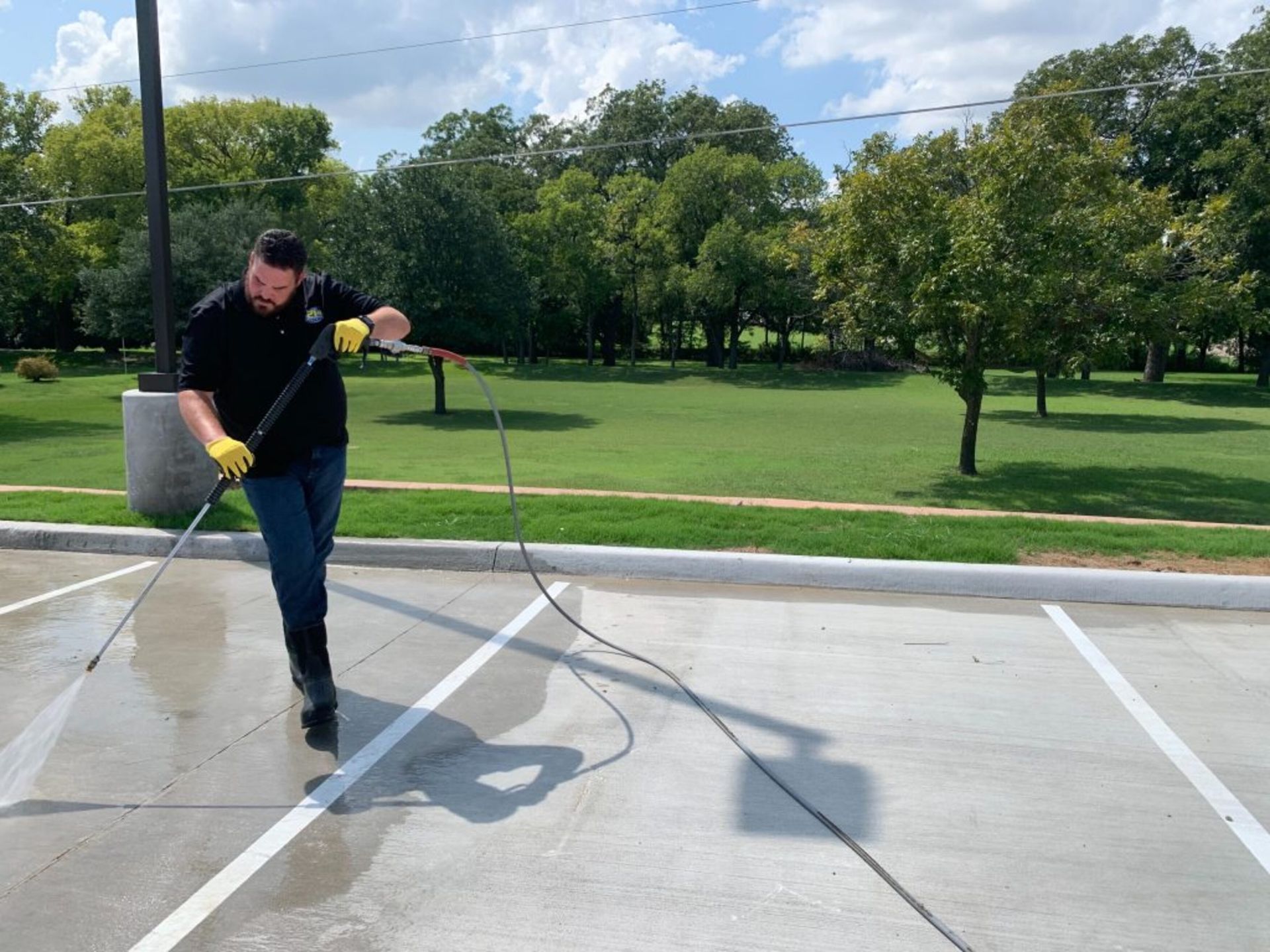 A person in dark clothing power washes a concrete parking space; trees in the background under a blue sky.