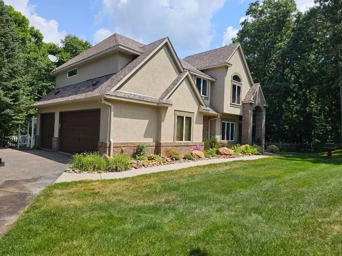 Two-story beige house with brown garage doors, green lawn, and trees under a cloudy sky.