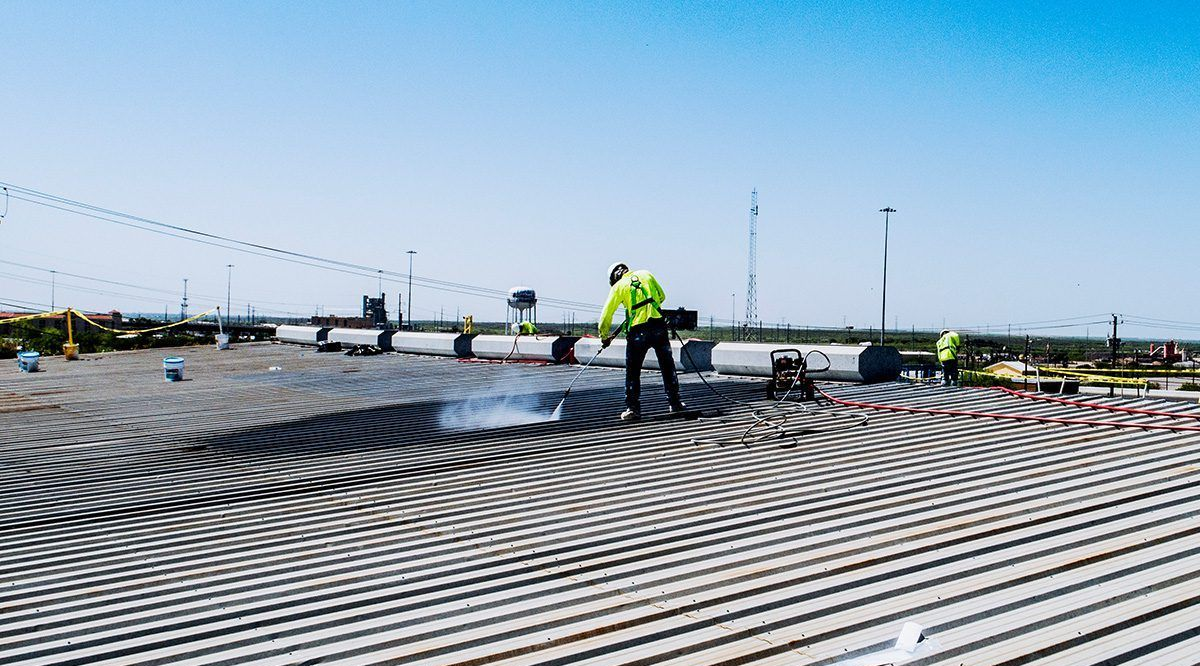 Workers cleaning a large, corrugated metal surface with pressure washers under a bright sky.