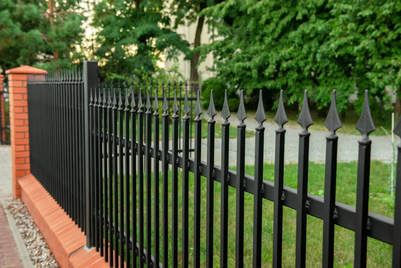 Black metal fence with pointed tops atop a brick base, separating a sidewalk from greenery.