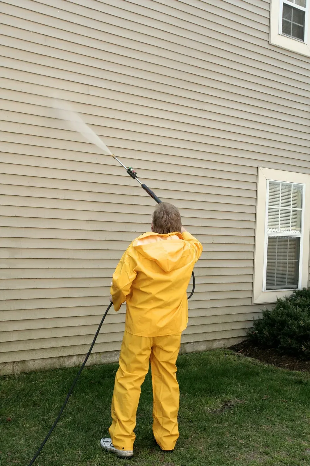 Person in yellow protective suit pressure washing siding of a house.