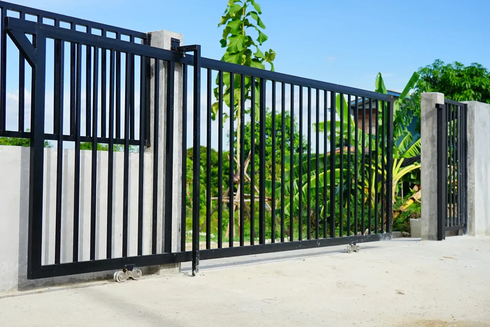 Black metal gate with vertical bars on a concrete wall, with greenery in the background.