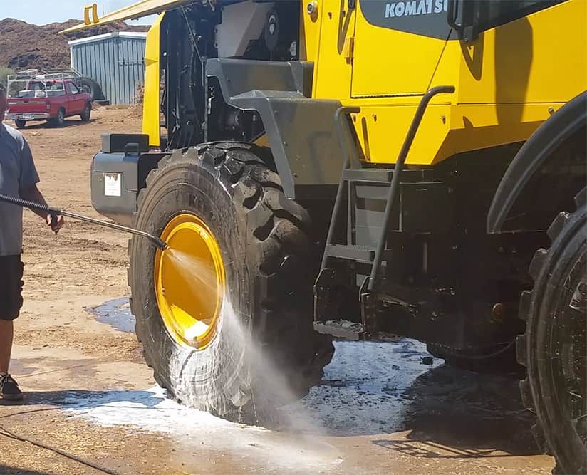 Person washing a large yellow construction vehicle's tire with a pressure washer outdoors.