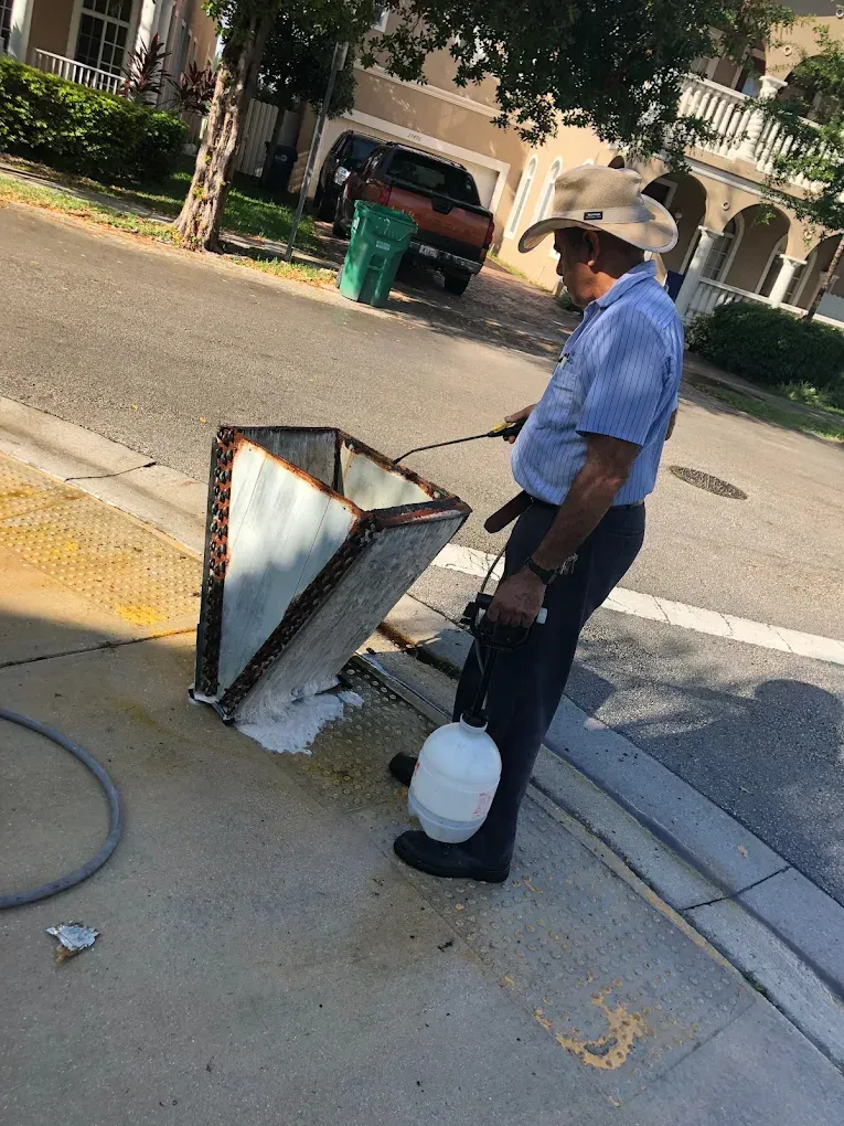 Man in a cowboy hat cleaning a metal box on a sidewalk with a hose and a bucket.