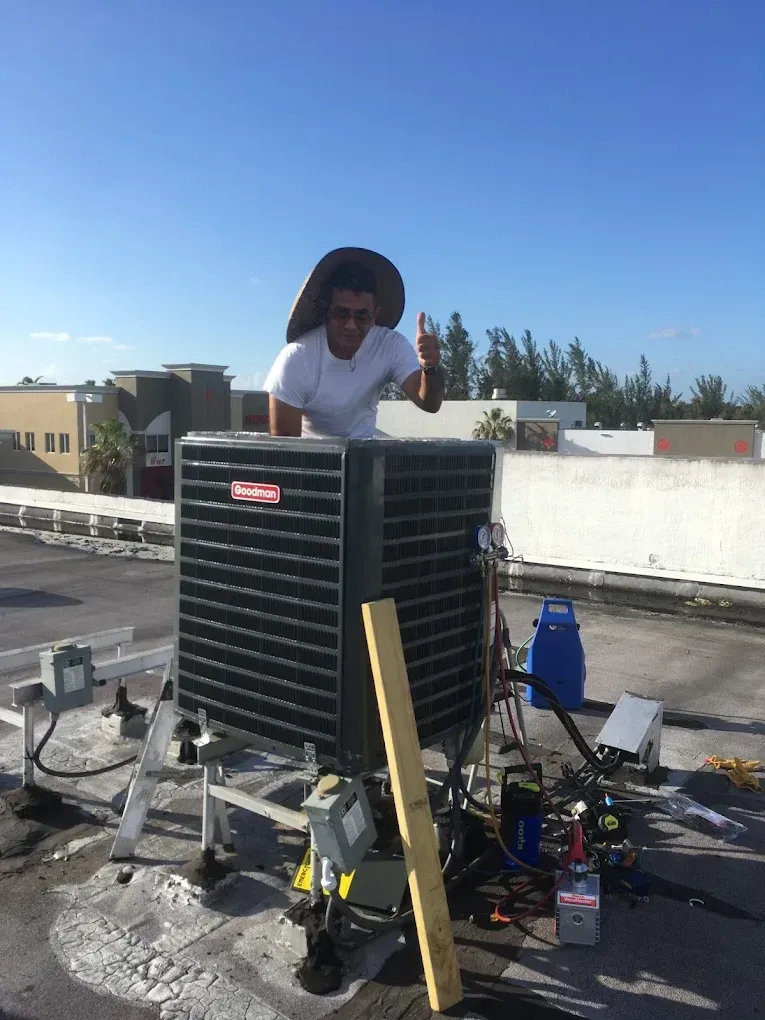 Person on rooftop beside air conditioner, giving thumbs-up. Blue sky, tools visible.