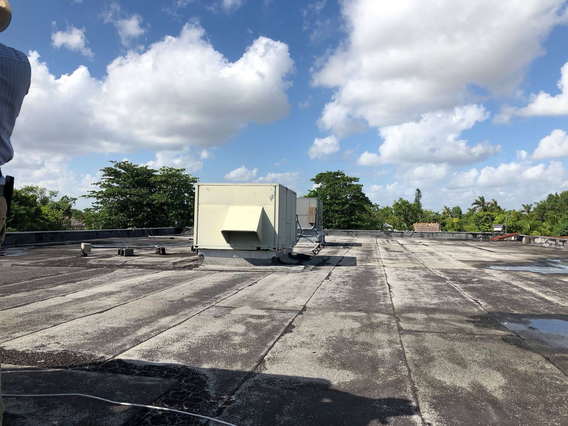 Flat, weathered roof with HVAC unit, trees, and cloudy blue sky.