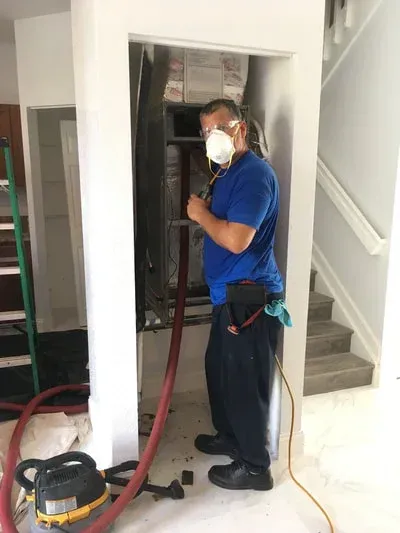 HVAC technician in a doorway, wearing a mask, working on ductwork, with a vacuum hose.