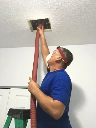 Person wearing a mask and headlamp, cleaning an air vent with a red hose.