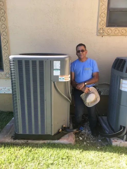Man stands beside an air conditioning unit outside a building. He holds a hat.