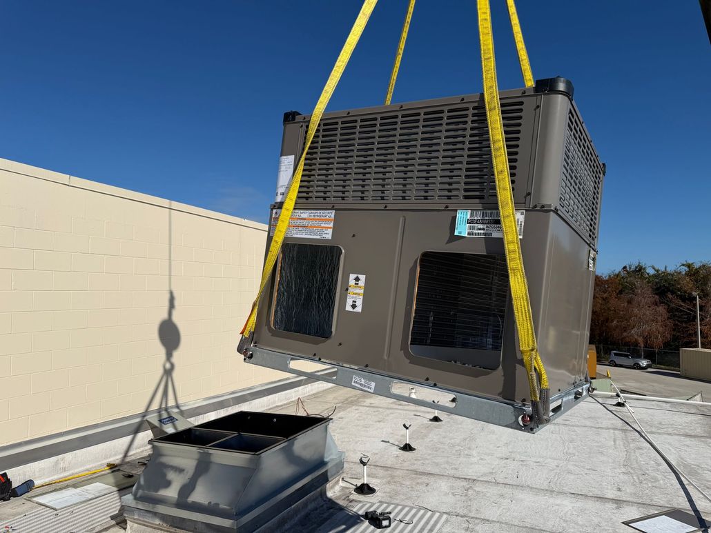 HVAC unit being lifted by yellow straps above a rooftop opening on a sunny day.