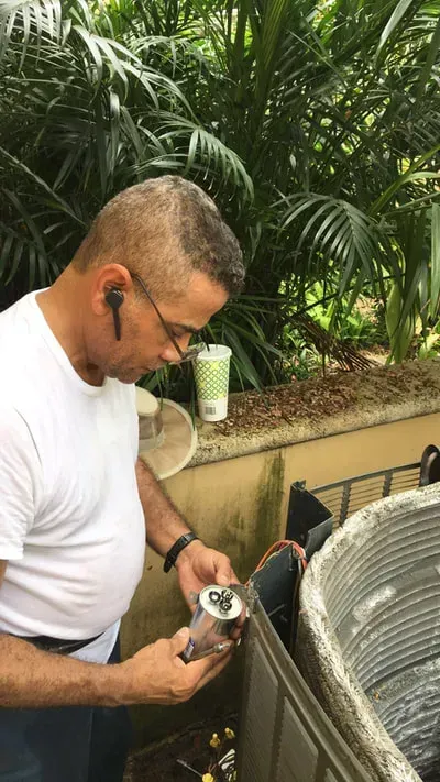 Man in white shirt repairs an AC unit outdoors; he holds a part and is wearing earbuds.