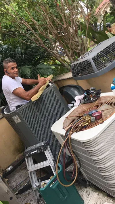 Man sitting in an air conditioning unit, smiling and giving a thumbs up, surrounded by equipment outdoors.
