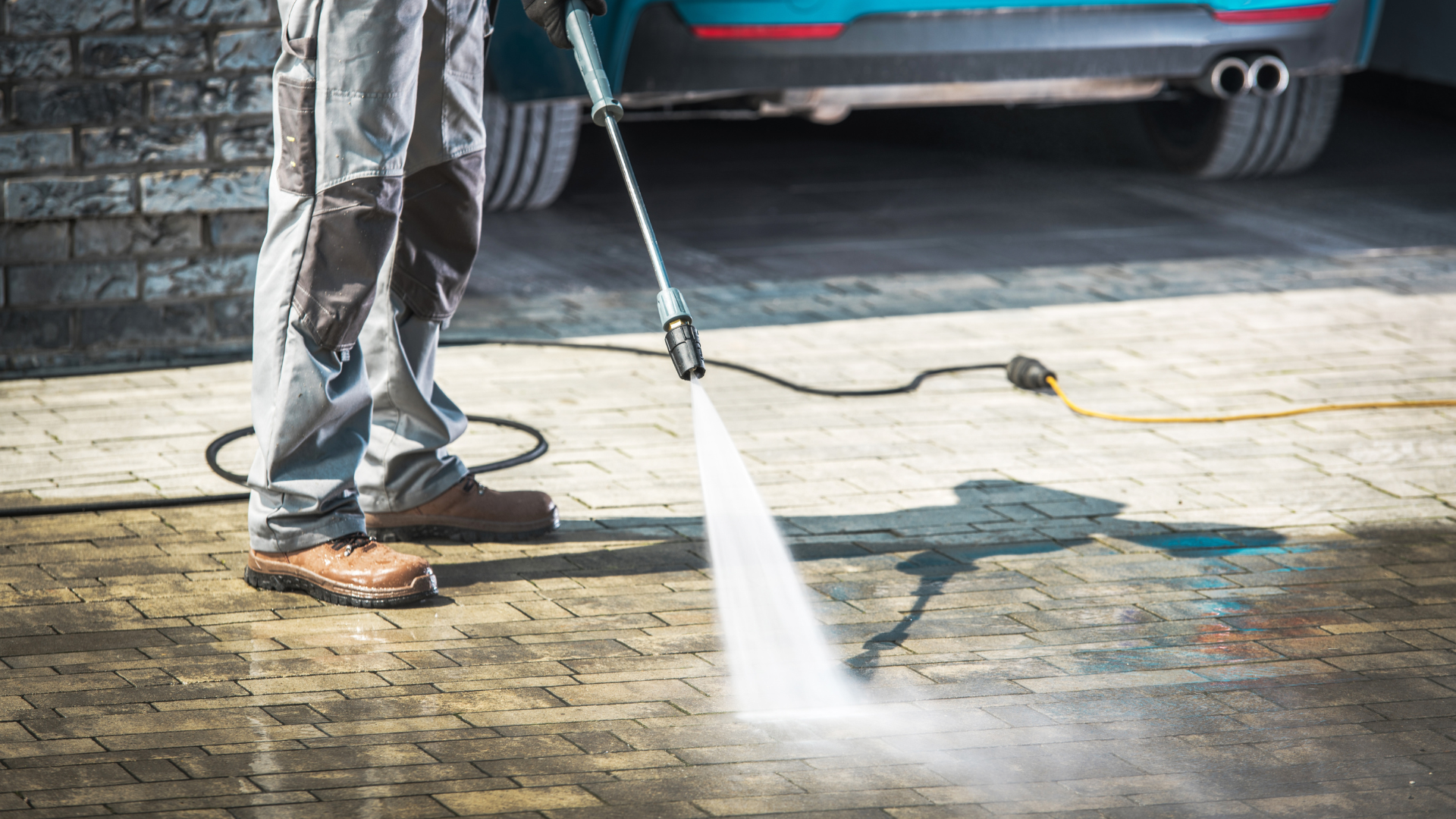 Person power washing a brick driveway next to a blue car.