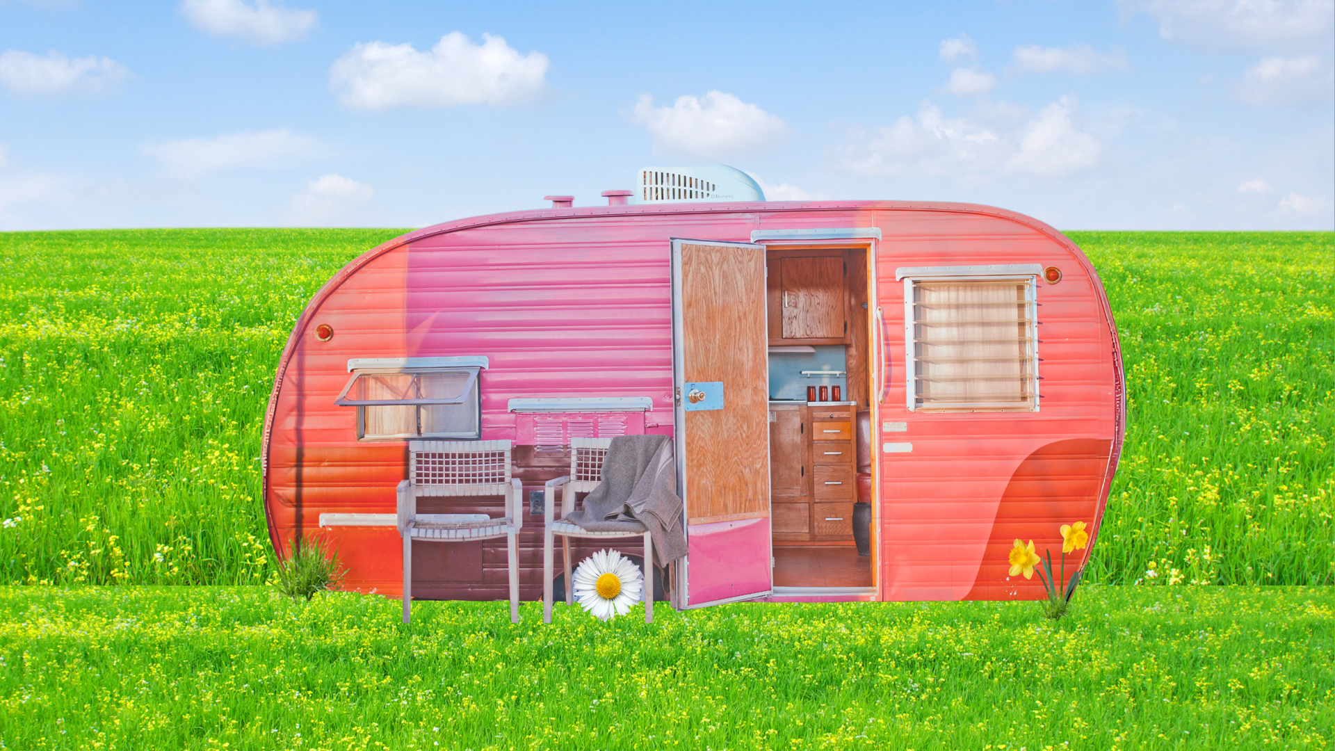 A vintage, pink and orange camper trailer sits in a vibrant, sunlit green field under a blue, cloudy sky.
