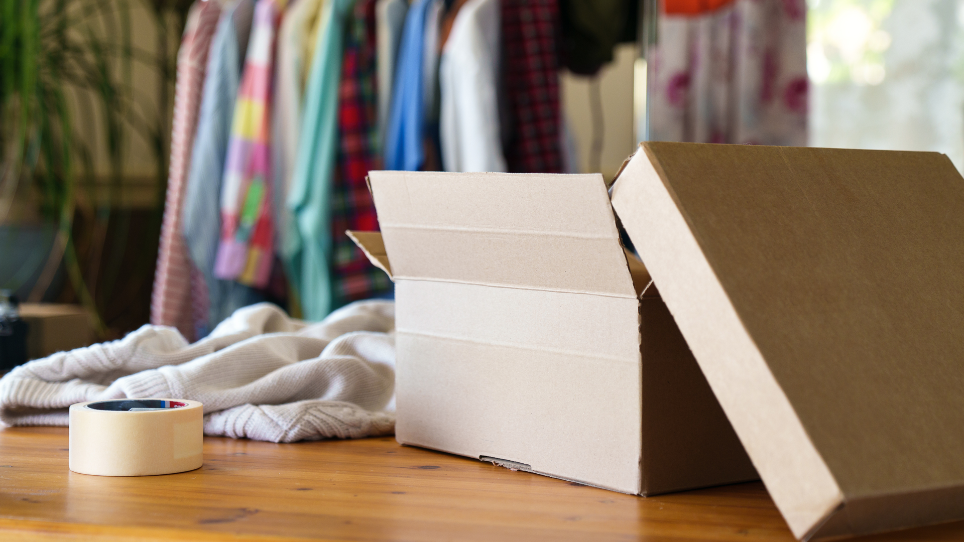 An open cardboard shipping box sits on a wooden table next to a roll of tape and folded clothing, with shirts on a rack.