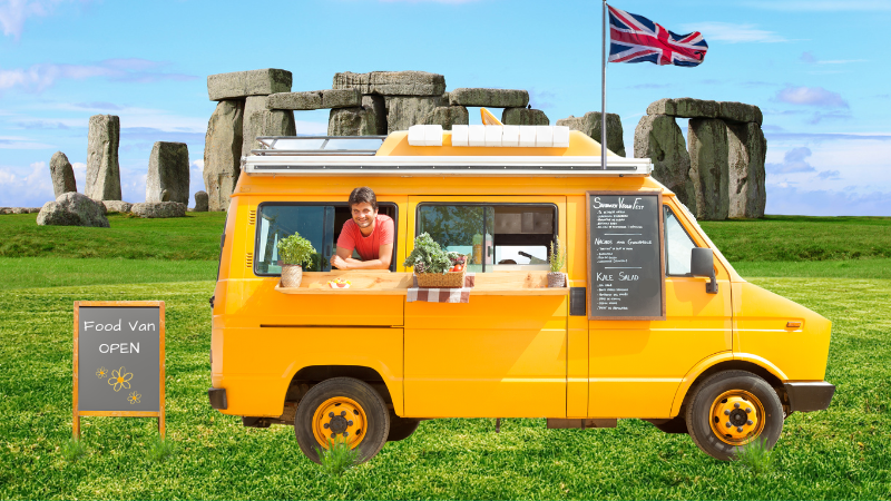 A person leans out of a bright yellow food van parked in a grassy field in front of Stonehenge with a Union Jack flag.