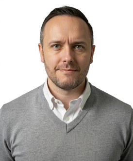 A person with short dark hair and a light beard wears a grey V-neck sweater over a white collared shirt against a white background.