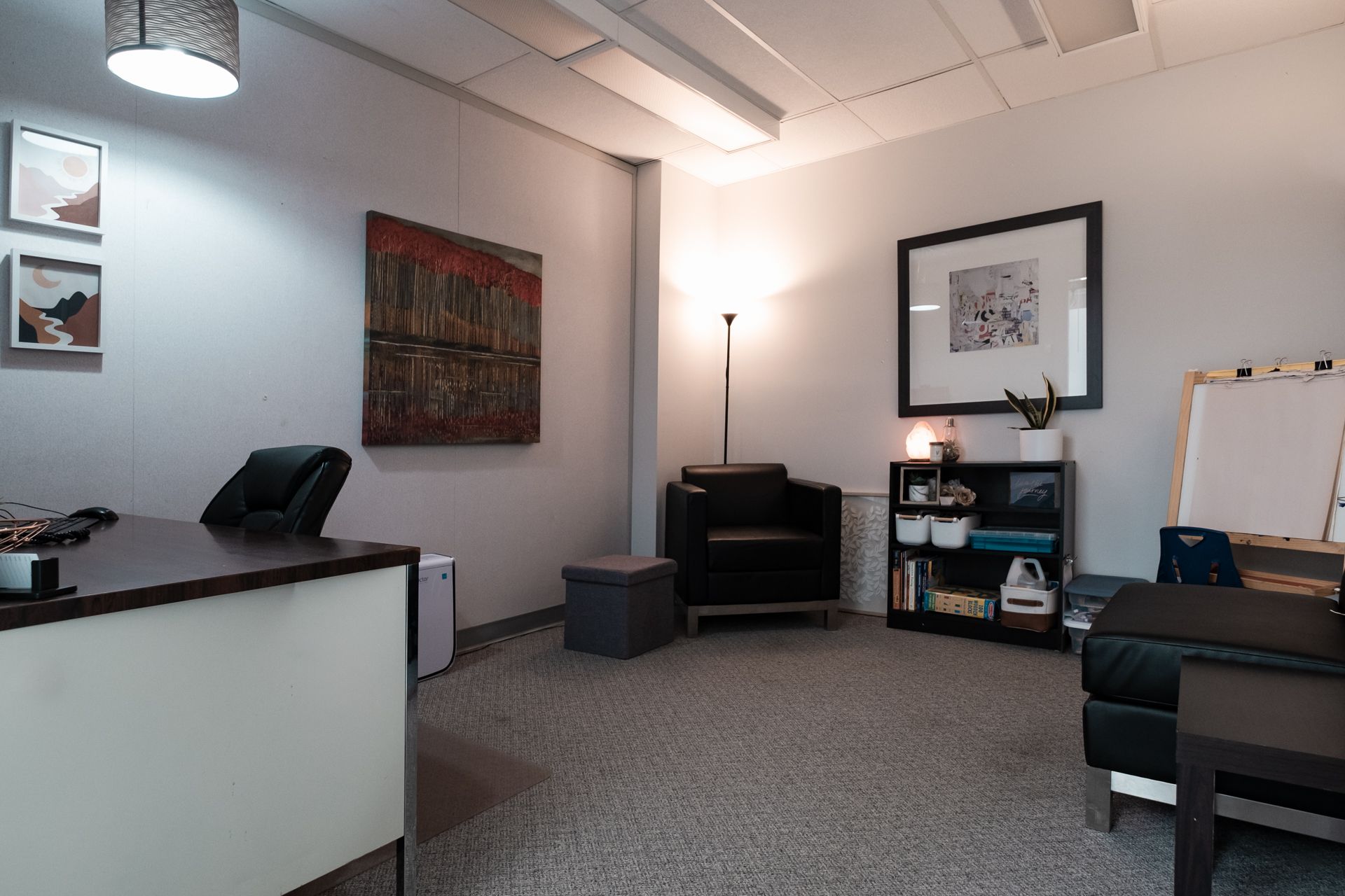 A living room with a desk , chairs , ottoman and paintings on the wall.