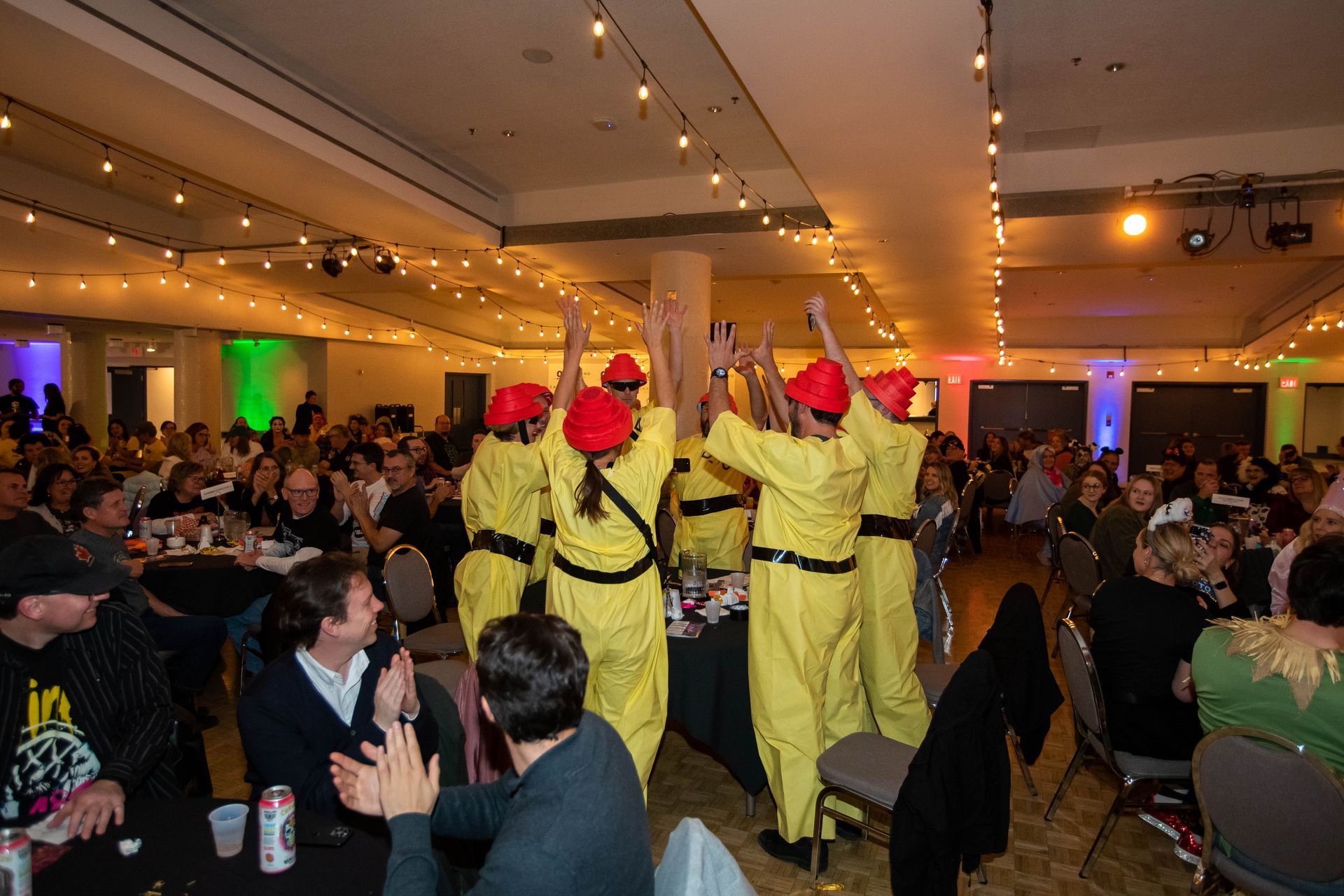 A group of people in yellow jumpsuits and red hats are dancing in a large room.