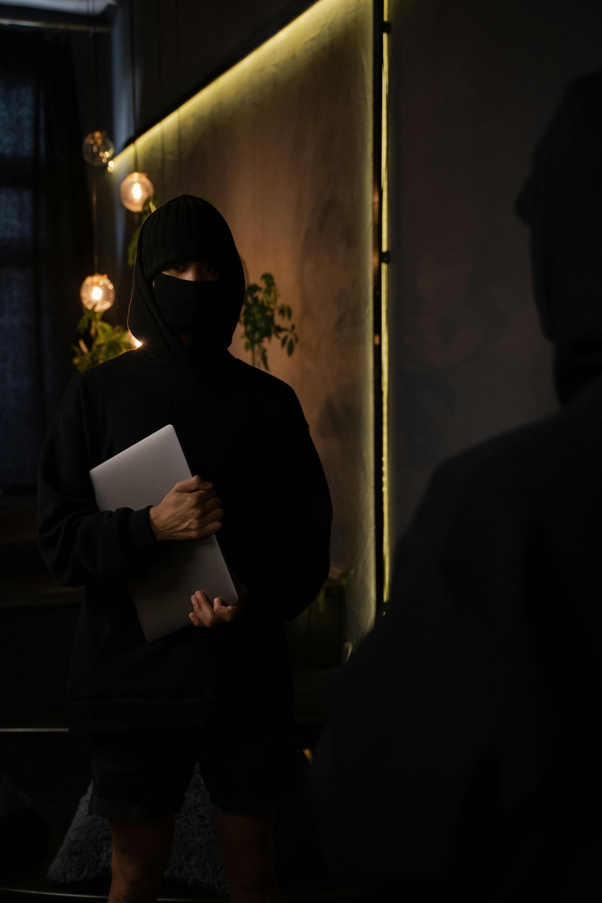 A man in a mask is holding a laptop in a dark room.