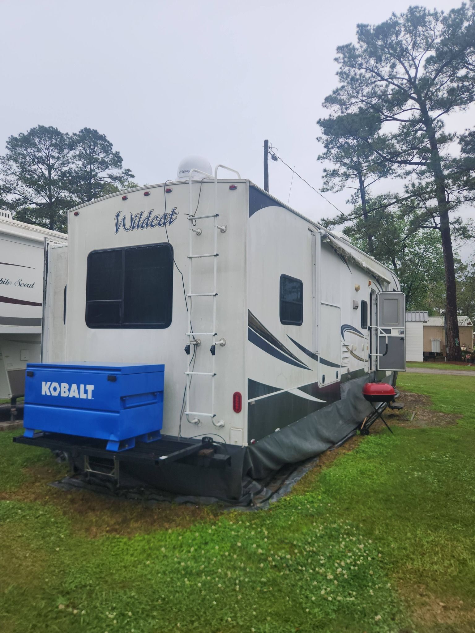 White Keystone RV with blue cargo box parked on grass beside another camper under cloudy sky