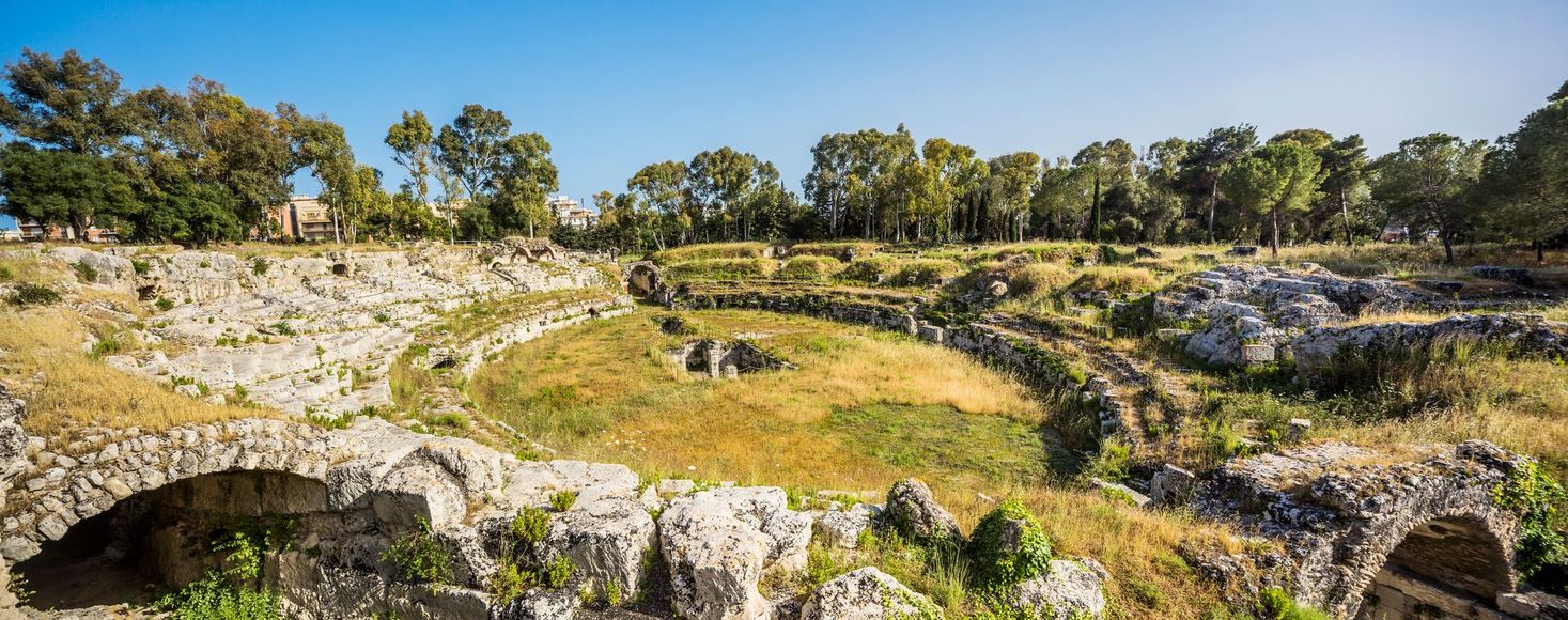 Una mano che mescola un cappuccino schiumoso in una tazza nera Mado, un croissant e dei bicchieri d'acqua su un tavolo.