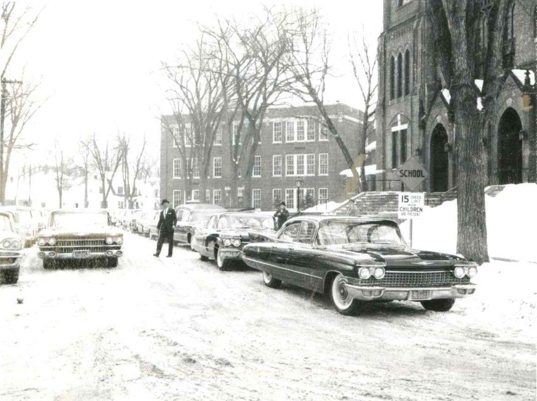 A black and white photo of cars parked on a snowy street in front of a church.